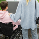 Une personne est dans un fauteuil roulant, une personne, debout, a posé sa main sur son épaule.